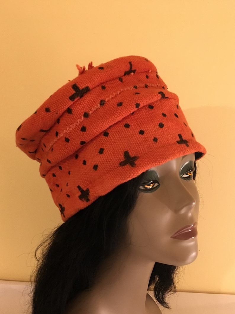 May include: An orange knit hat with black cross and dot patterns. The hat has a folded design and a small fringe detail. The hat is displayed on a mannequin head with black hair.