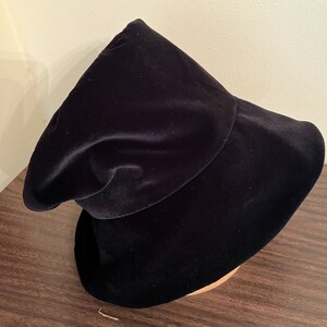 May include: A black velvet hat with a wide brim and a rounded crown. The hat is displayed on a wooden hat block. The hat's fabric has a soft, luxurious appearance, and the overall design is elegant and classic.