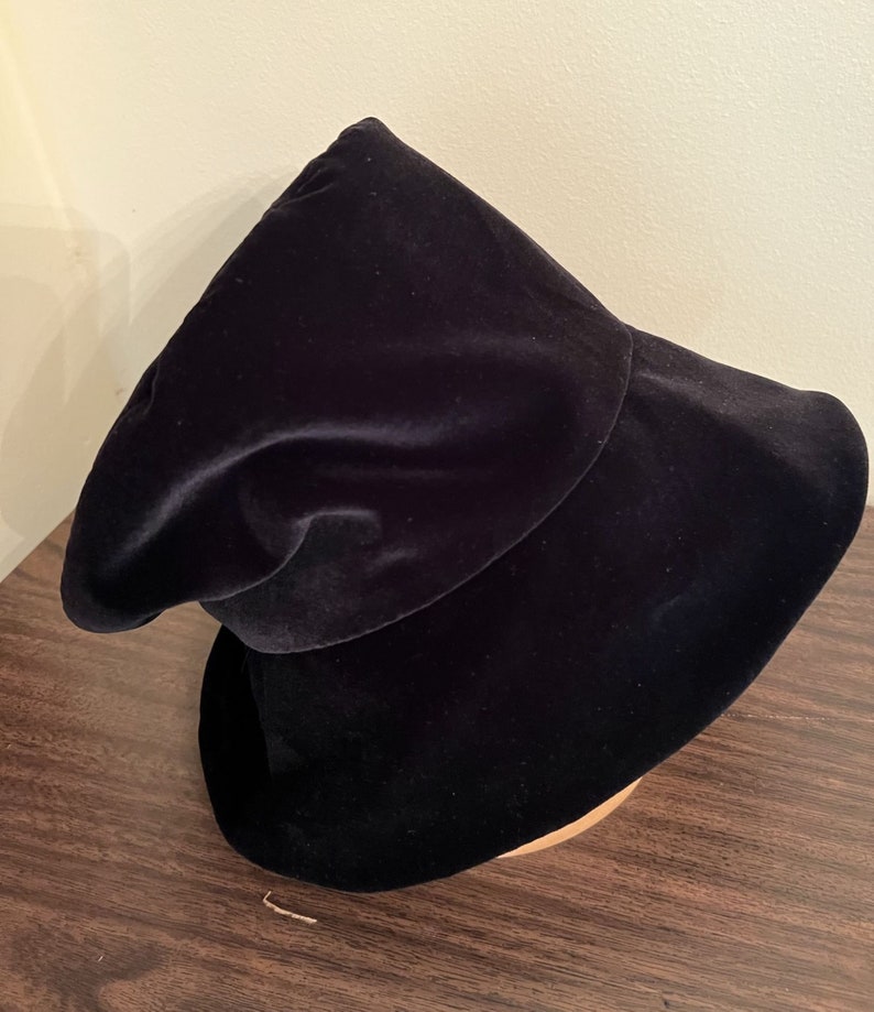 May include: A black velvet hat with a wide brim and a rounded crown. The hat is displayed on a wooden hat block. The hat's fabric has a soft, luxurious appearance, and the overall design is elegant and classic.