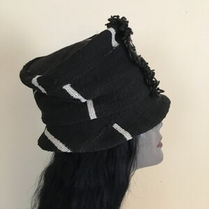 May include: Black hat with white stripes and a fringed edge. The hat has a structured, layered design. The hat is made of a woven material. The hat is a fashion accessory.