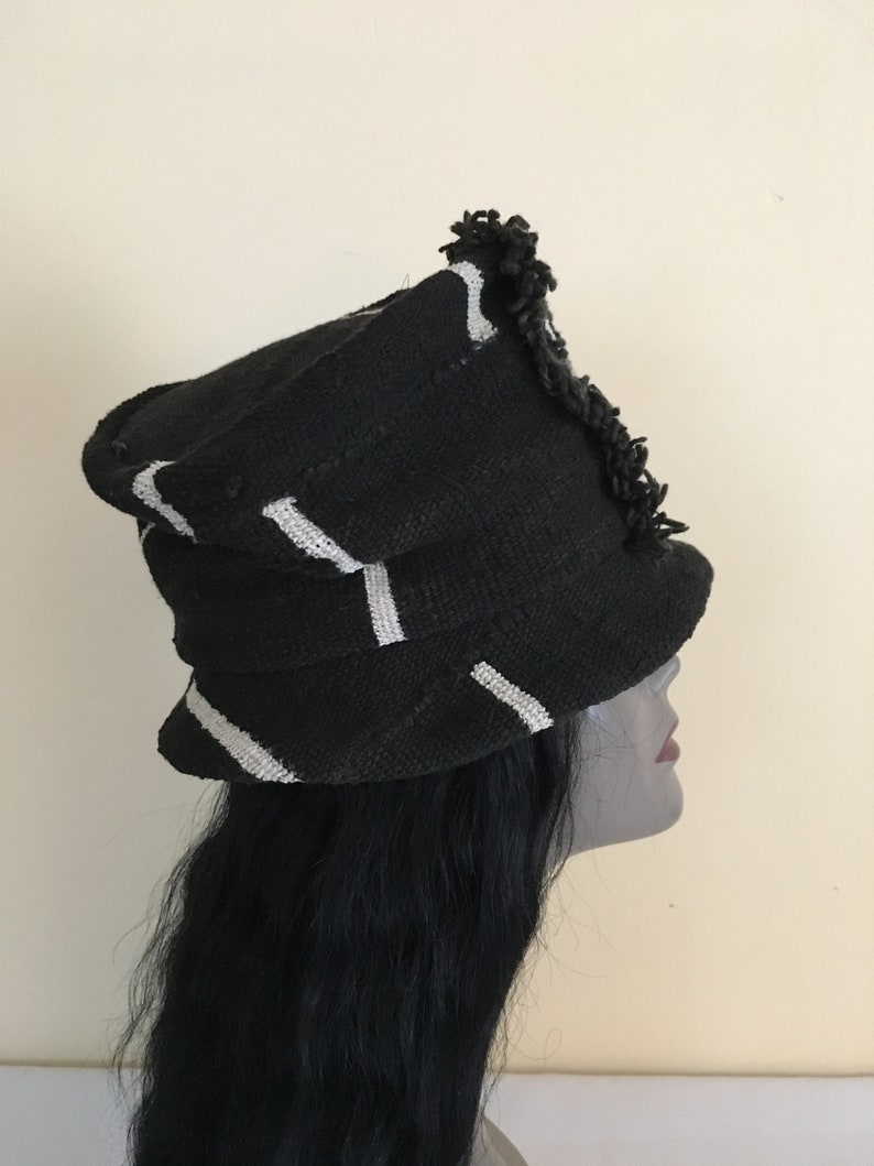 May include: Black hat with white stripes and a fringed edge. The hat has a structured, layered design. The hat is made of a woven material. The hat is a fashion accessory.