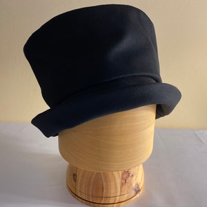 May include: A black top hat with a rounded crown and a wide, flat brim. The hat is made of a dark, possibly felt, material. It sits on a wooden hat block, showcasing its shape and structure. The hat is a classic accessory.