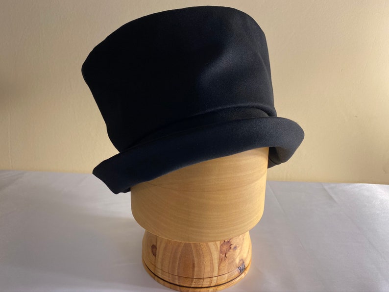 May include: A black top hat with a rounded crown and a wide, flat brim. The hat is made of a dark, possibly felt, material. It sits on a wooden hat block, showcasing its shape and structure. The hat is a classic accessory.
