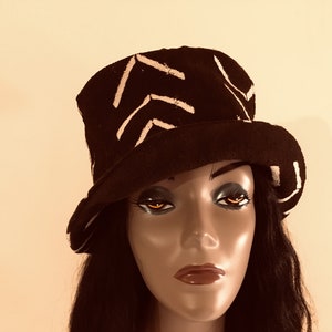 May include: A black hat with a white chevron pattern. The hat has a wide brim and a cylindrical crown. The hat is displayed on a mannequin head with dark hair and makeup. The background is a plain, neutral colour.