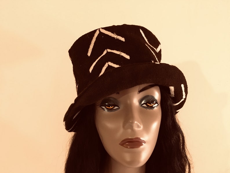 May include: A black hat with a white chevron pattern. The hat has a wide brim and a cylindrical crown. The hat is displayed on a mannequin head with dark hair and makeup. The background is a plain, neutral colour.