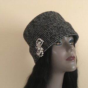 May include: A gray and black striped cloche hat with a decorative rhinestone brooch. The hat has a rounded crown and a small brim. The brooch is a cluster of clear rhinestones in a floral design. A stylish accessory.