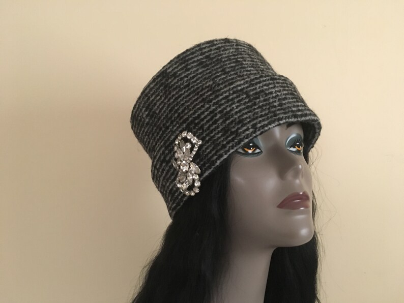 May include: A gray and black striped cloche hat with a decorative rhinestone brooch. The hat has a rounded crown and a small brim. The brooch is a cluster of clear rhinestones in a floral design. A stylish accessory.