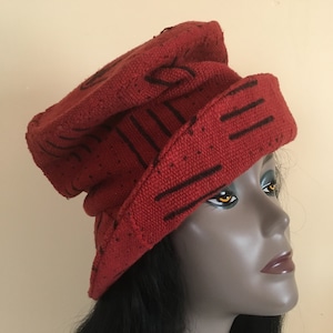 K&ouml;nnte beinhalten: Ein roter Strickhut mit schwarzen linearen und geschwungenen Designs. Der Hut wird auf einem Schaufensterkopf getragen. Der Hut hat eine Krempe und eine abgerundete Oberseite. Der Hut besteht aus einem strukturierten Material.