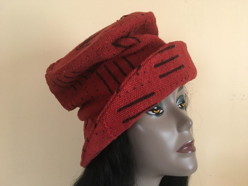 K&ouml;nnte beinhalten: Ein roter Strickhut mit schwarzen linearen und geschwungenen Designs. Der Hut wird auf einem Schaufensterkopf getragen. Der Hut hat eine Krempe und eine abgerundete Oberseite. Der Hut besteht aus einem strukturierten Material.