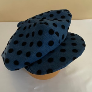 May include: A blue beret with black polka dots. The hat is made of a soft fabric and is folded over. The beret is sitting on a wooden form.