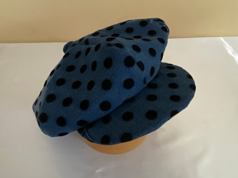 May include: A blue beret with black polka dots. The hat is made of a soft fabric and is folded over. The beret is sitting on a wooden form.