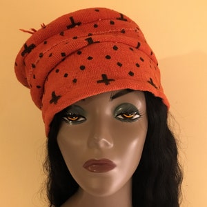 May include: An orange turban-style head covering with a repeating pattern of black plus signs and dots. The fabric has a textured appearance. The head covering is worn by a mannequin with black hair.