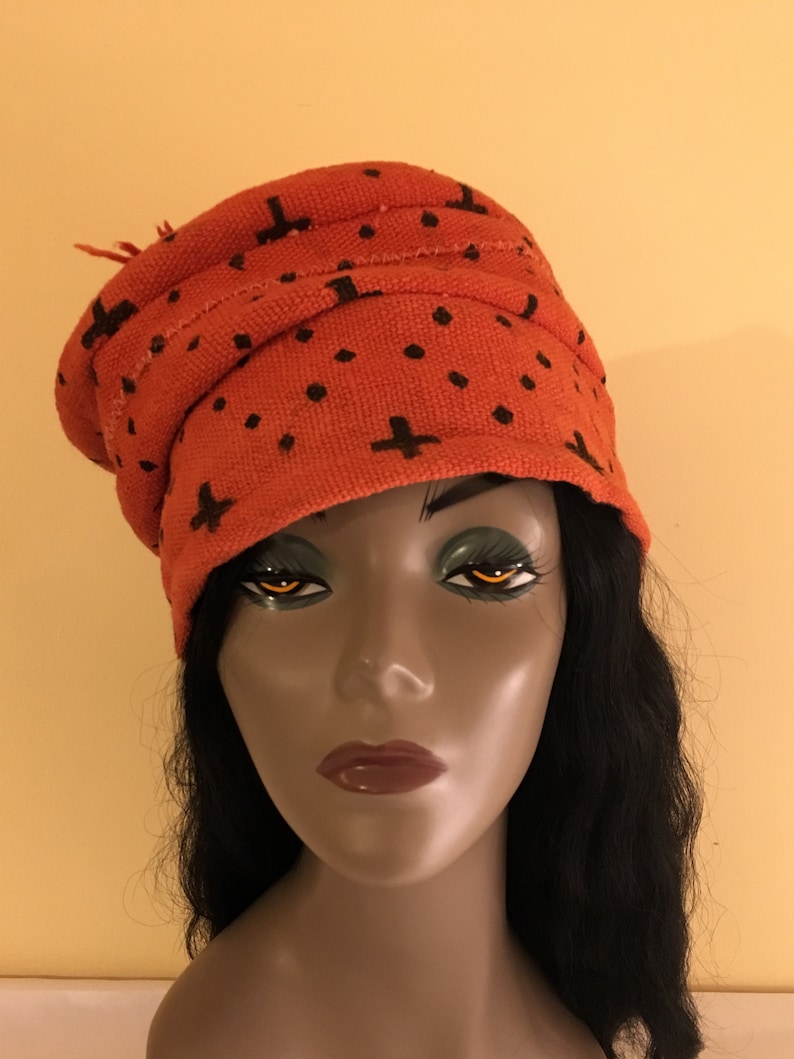 May include: An orange turban-style head covering with a repeating pattern of black plus signs and dots. The fabric has a textured appearance. The head covering is worn by a mannequin with black hair.