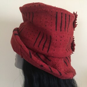 K&ouml;nnte beinhalten: Ein roter Hut mit schwarzen vertikalen Linienmustern und kleinen gepunkteten Details. Der Hut hat eine gerollte Krempe und einen Fransenakzent. Der Hut ist aus einem gewebten Stoff gefertigt.