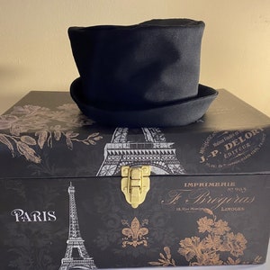 May include: A black top hat with a rounded crown and a wide brim sits atop a decorative black box. The box features Parisian imagery, including the Eiffel Tower and floral designs, with gold accents and text.