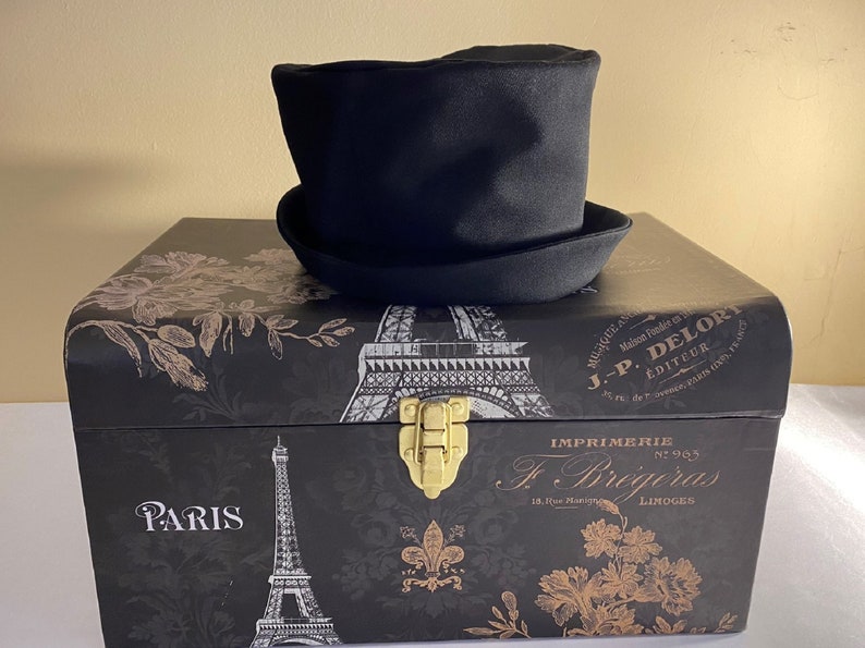 May include: A black top hat with a rounded crown and a wide brim sits atop a decorative black box. The box features Parisian imagery, including the Eiffel Tower and floral designs, with gold accents and text.