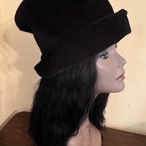 May include: A black hat with a wide brim and a tall, slightly conical crown. The hat is displayed on a mannequin head with long, black wavy hair. The hat's design features a folded brim, adding a touch of elegance to the overall look.