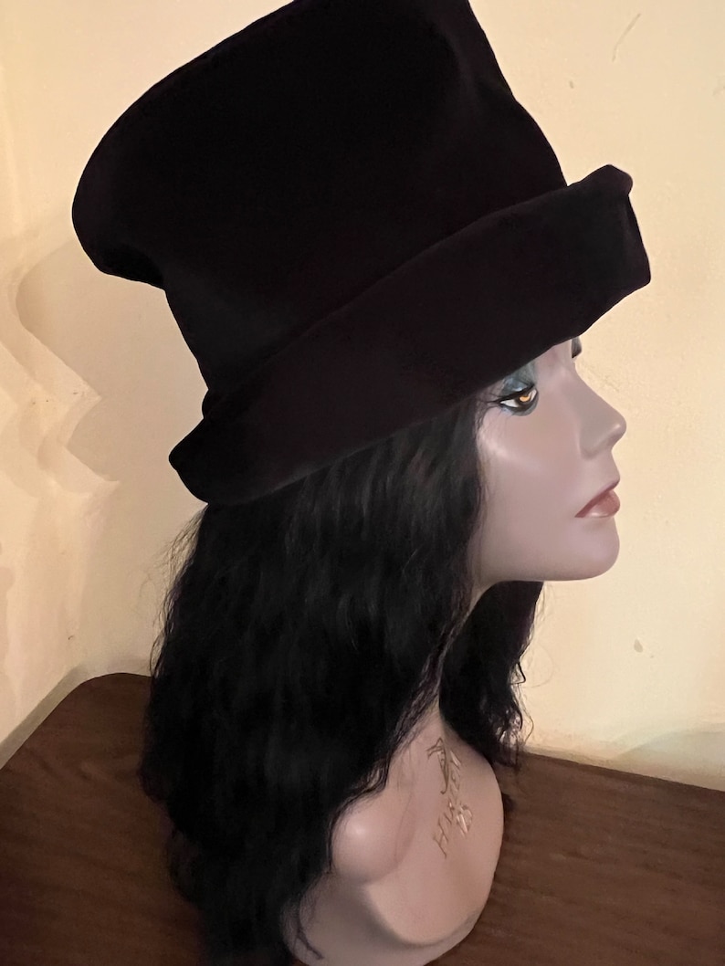 May include: A black hat with a wide brim and a tall, slightly conical crown. The hat is displayed on a mannequin head with long, black wavy hair. The hat's design features a folded brim, adding a touch of elegance to the overall look.