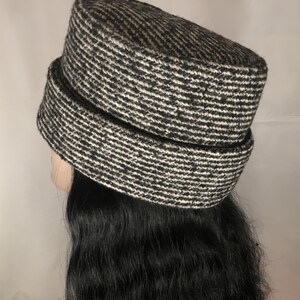 May include: A black and white patterned hat with a cylindrical crown and a wide brim. The hat has a textured appearance and is made of a woven fabric. The hat is shown on a mannequin head with long, black hair.