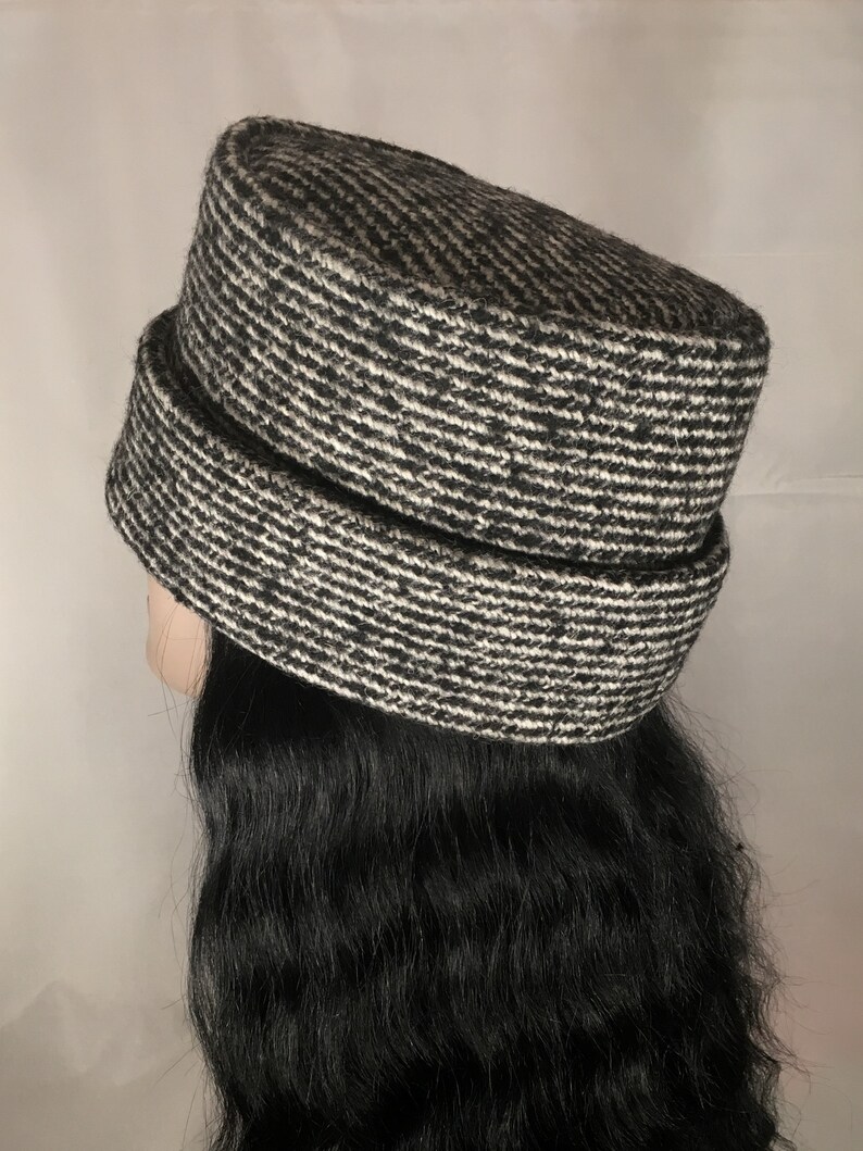 May include: A black and white patterned hat with a cylindrical crown and a wide brim. The hat has a textured appearance and is made of a woven fabric. The hat is shown on a mannequin head with long, black hair.