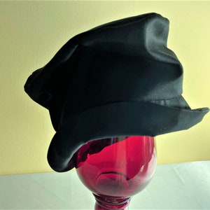 May include: A black hat with a unique, slightly asymmetrical shape. The hat has a wide brim and a textured appearance. It is displayed on top of a red glass object, creating a striking contrast against a yellow background.