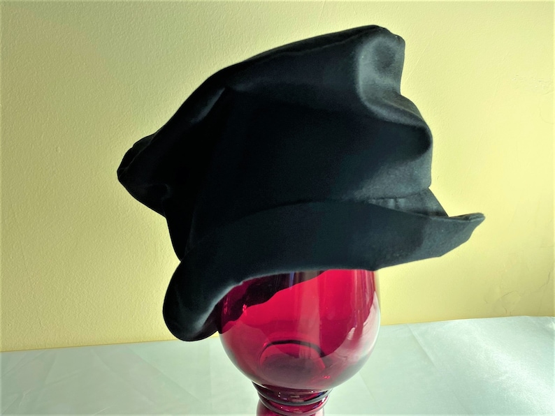 May include: A black hat with a unique, slightly asymmetrical shape. The hat has a wide brim and a textured appearance. It is displayed on top of a red glass object, creating a striking contrast against a yellow background.