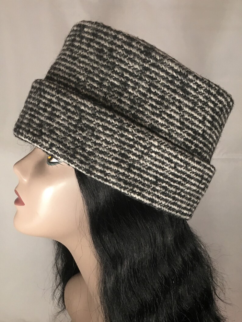 May include: A black and white patterned hat with a rectangular crown and a folded brim. The hat is made of a textured fabric and is displayed on a mannequin head. The hat's design features a diagonal stripe pattern.