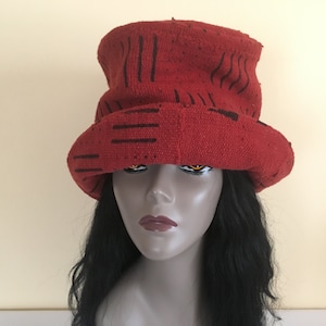 Puede incluir: Un sombrero rojo con diseños lineales negros. El sombrero tiene un ala ancha y una apariencia ligeramente texturizada. El sombrero se muestra en una cabeza de maniquí con cabello negro y un fondo neutro.