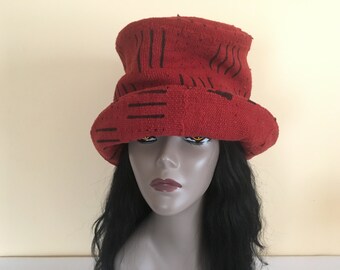 May include: A red bucket hat with black linear designs. The hat has a wide brim and a slightly textured appearance. The hat is displayed on a mannequin head with black hair and a neutral background.