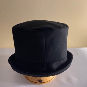 May include: A black top hat with a rounded crown and a wide brim. The hat is made of a dark fabric and sits on a wooden hat stand. The hat is a classic formal accessory.