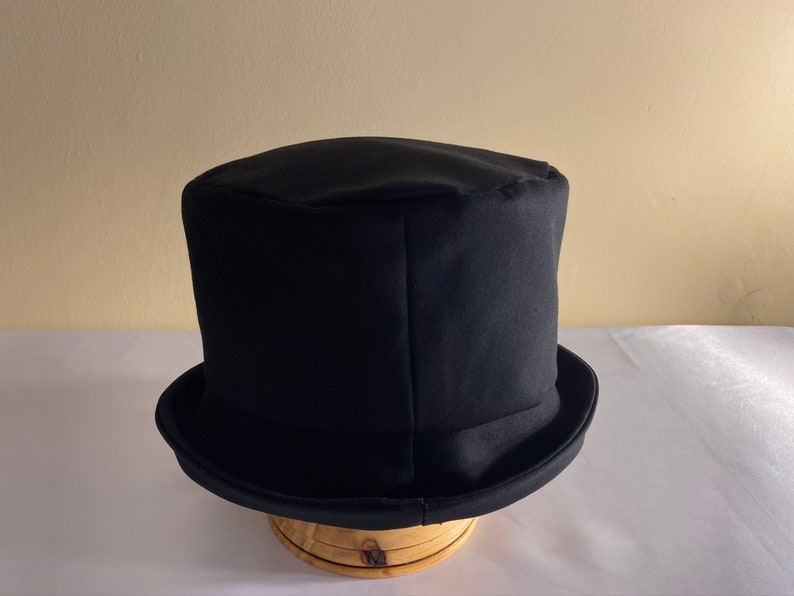 May include: A black top hat with a rounded crown and a wide brim. The hat is made of a dark fabric and sits on a wooden hat stand. The hat is a classic formal accessory.