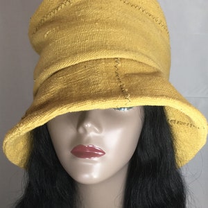 May include: A mustard yellow bucket hat made of textured fabric. The hat features a slightly slouched crown and a wide brim. Visible stitching accents the seams. The hat is displayed on a mannequin head with dark hair.