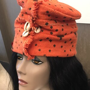 May include: An orange fabric hat with black polka dots and fringe trim. The hat is adorned with three white seashells. The hat is displayed on a mannequin head with black hair.