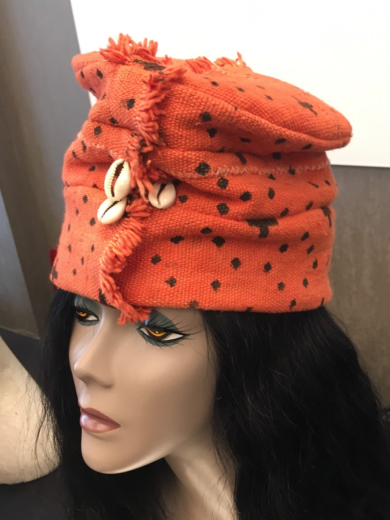 May include: An orange fabric hat with black polka dots and fringe trim. The hat is adorned with three white seashells. The hat is displayed on a mannequin head with black hair.