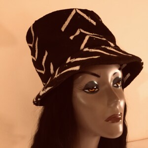 May include: Black bucket hat with a white zigzag design. The hat is shown on a mannequin head. The hat has a short brim and a rounded crown. The hat is made of a textured material.