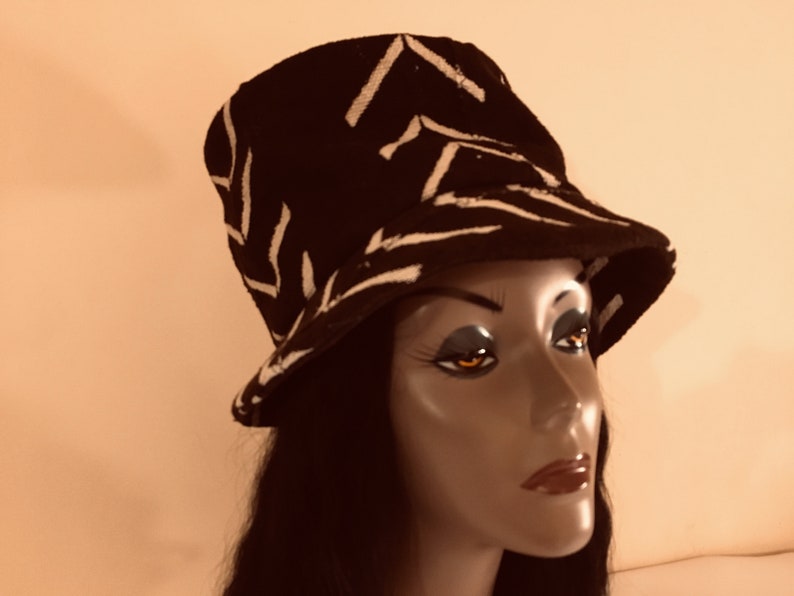 May include: Black bucket hat with a white zigzag design. The hat is shown on a mannequin head. The hat has a short brim and a rounded crown. The hat is made of a textured material.