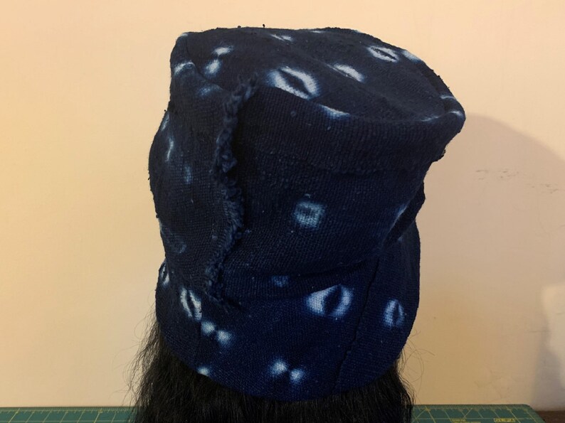 Puede incluir: Un sombrero azul oscuro con ala, con un patr&oacute;n de dise&ntilde;os blancos en forma de ojo. El sombrero tiene un borde desgastado, lo que le da un aspecto artesanal. El sombrero est&aacute; hecho de una tela texturizada.