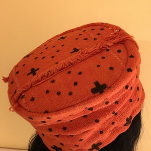 May include: An orange fabric hat with a black cross and dot pattern. The hat has a rounded shape with a fringed edge. The hat is worn on a person's head.