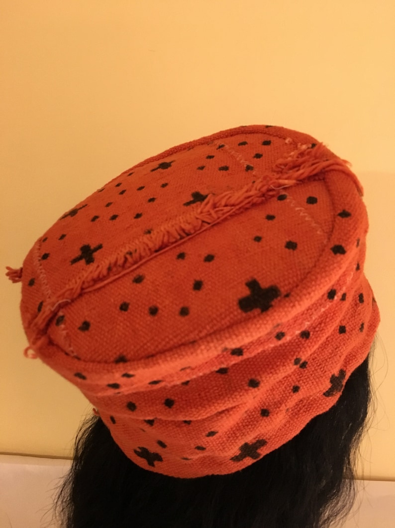 May include: An orange fabric hat with a black cross and dot pattern. The hat has a rounded shape with a fringed edge. The hat is worn on a person's head.