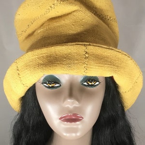 Puede incluir: Un sombrero amarillo mostaza con ala ancha y copa redondeada. El sombrero está hecho de una tela texturizada y presenta costuras visibles a lo largo de las costuras. El sombrero se exhibe en una cabeza de maniquí con cabello negro y rasgos pintados.