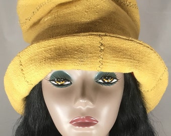 May include: A mustard yellow hat with a wide brim and a rounded crown. The hat is made of a textured fabric and features visible stitching along the seams. The hat is displayed on a mannequin head with black hair and painted features.