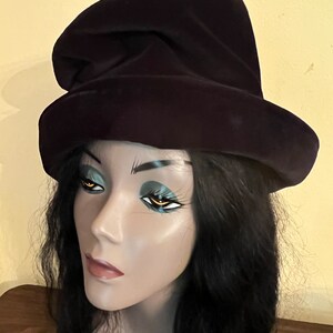 May include: A dark purple velvet hat with a wide brim and a tall, pointed crown. The hat is displayed on a mannequin head with black hair and dramatic eye makeup. The background is a light yellow color.