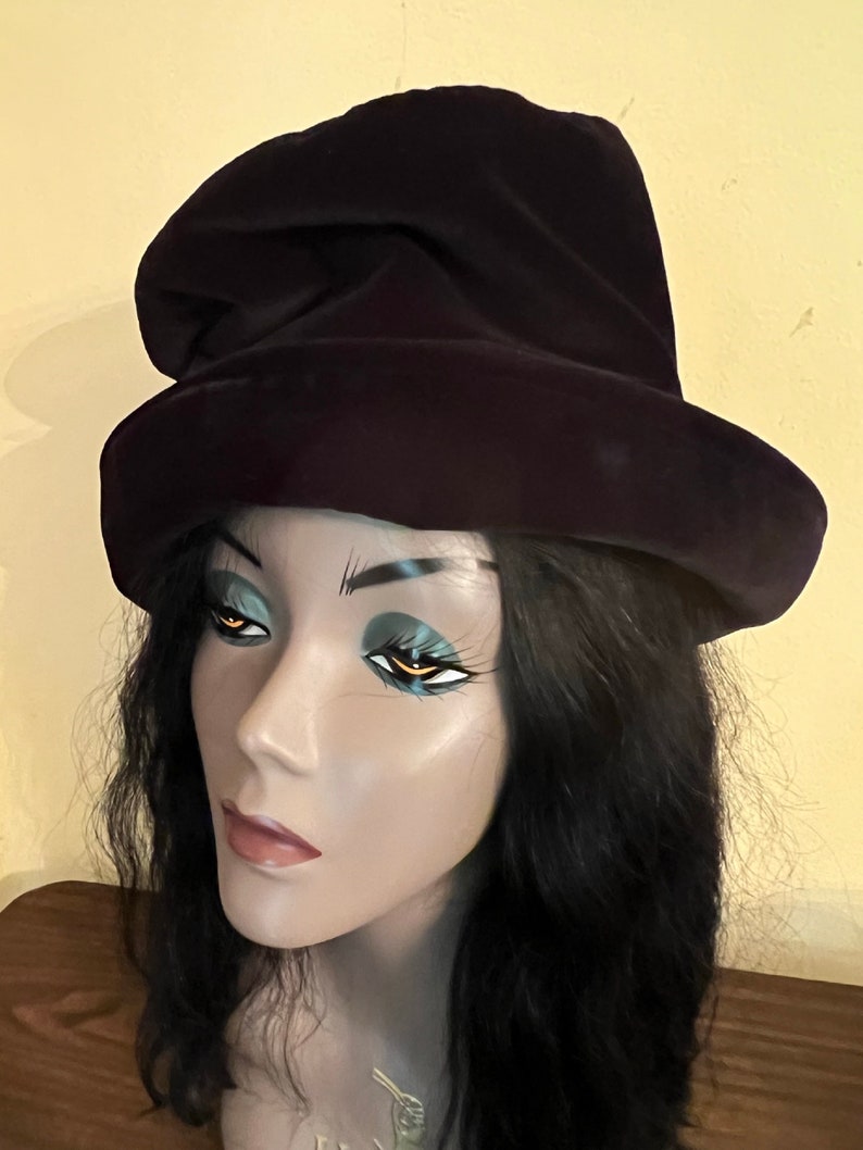 May include: A dark purple velvet hat with a wide brim and a tall, pointed crown. The hat is displayed on a mannequin head with black hair and dramatic eye makeup. The background is a light yellow color.