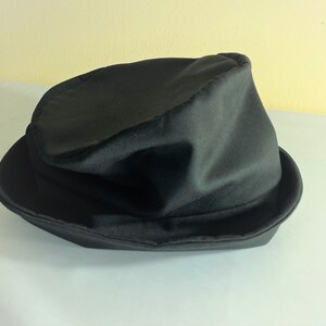 May include: A black fedora hat with a slightly crumpled crown and a wide brim. The hat is made of a soft, possibly felt-like material, and sits on a white surface. The hat's dark color suggests a classic, versatile accessory.