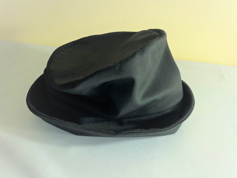 May include: A black fedora hat with a slightly crumpled crown and a wide brim. The hat is made of a soft, possibly felt-like material, and sits on a white surface. The hat's dark color suggests a classic, versatile accessory.