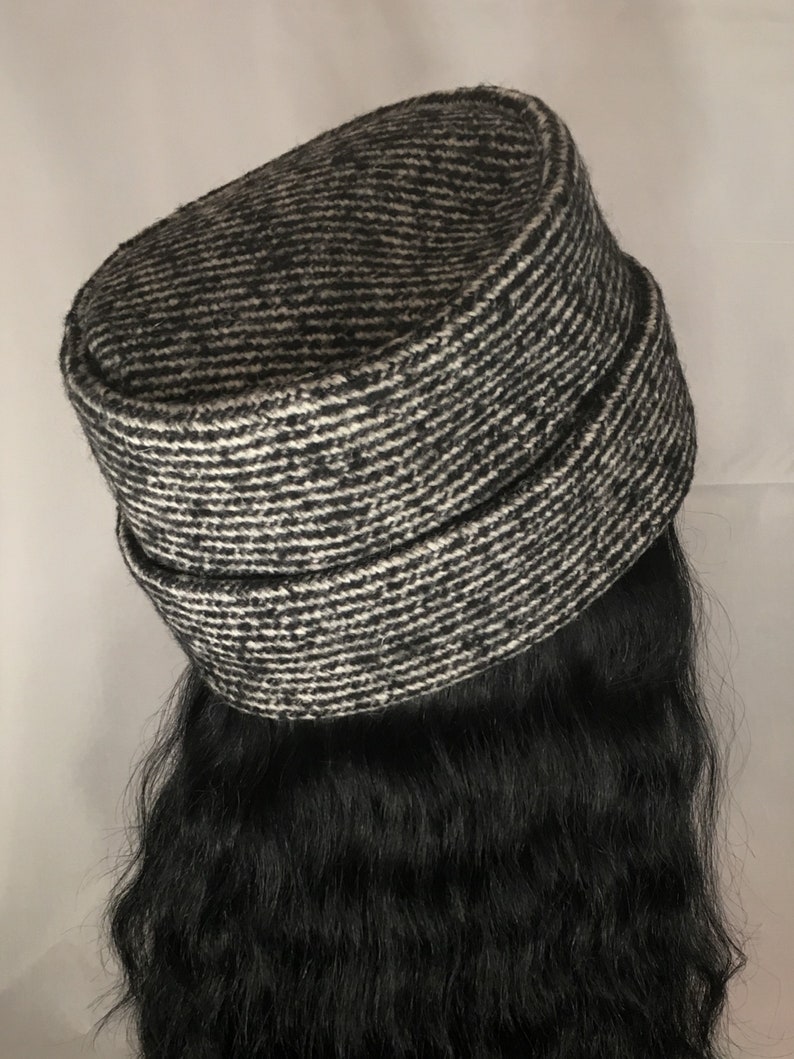 May include: A black and white tweed hat with a horizontal striped pattern. The hat has a rounded top and a band around the base. The hat is a neutral color and can be worn with many outfits.