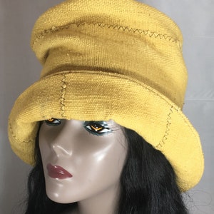 May include: A yellow bucket hat with a wide brim. The hat is made of a textured fabric and features decorative stitching along the seams. The hat is displayed on a mannequin head with black hair.