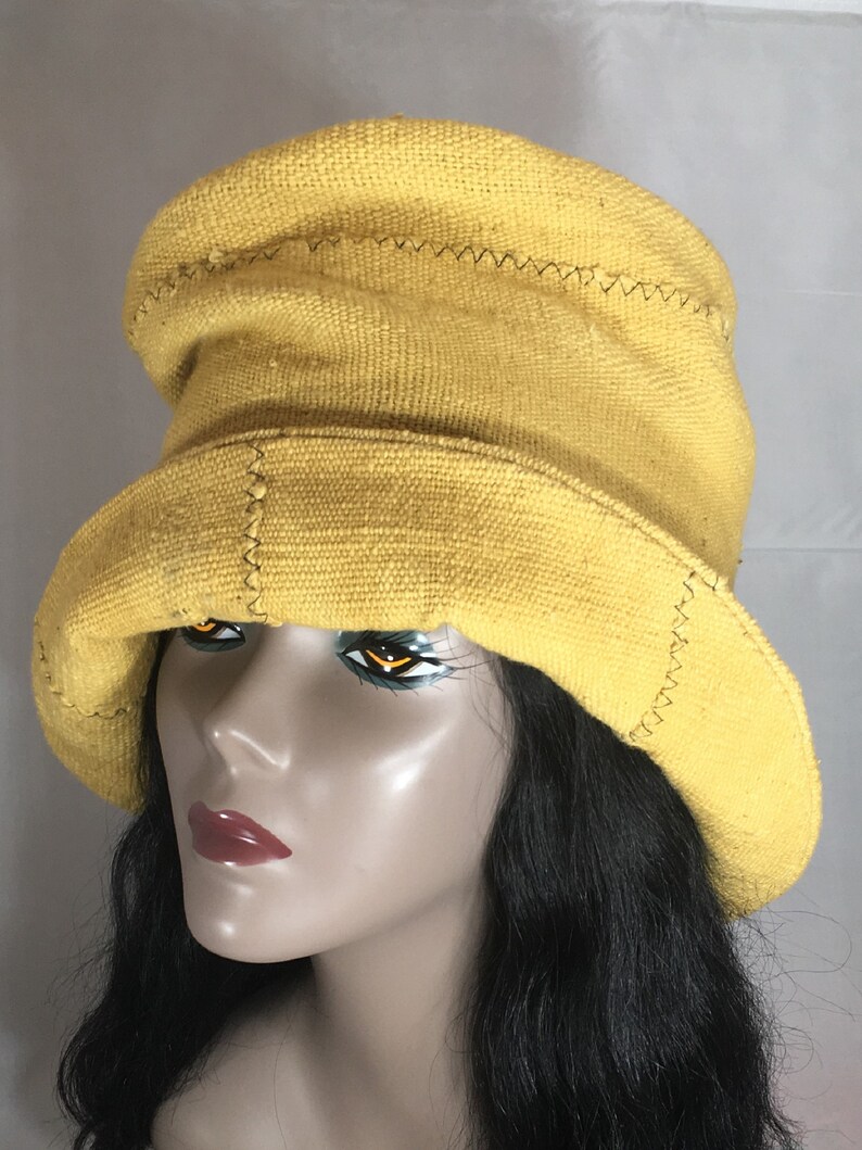 May include: A yellow bucket hat with a wide brim. The hat is made of a textured fabric and features decorative stitching along the seams. The hat is displayed on a mannequin head with black hair.
