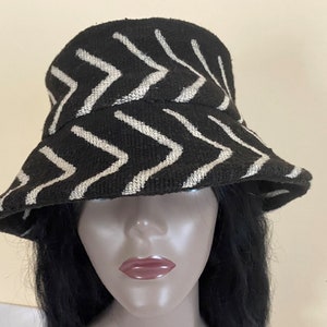 May include: Black bucket hat with a white zigzag pattern. The hat has a wide brim and a rounded crown. The fabric appears to be textured. This hat is a stylish accessory.