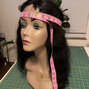 May include: A mannequin head with black hair and a pink measuring tape wrapped around the head. The tape is held in place with a metal clip. The mannequin has the words "HARLEM 125" on the neck.